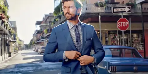 Man in a blue suit, perfect cocktail attire, stands on a street with a vintage car.
