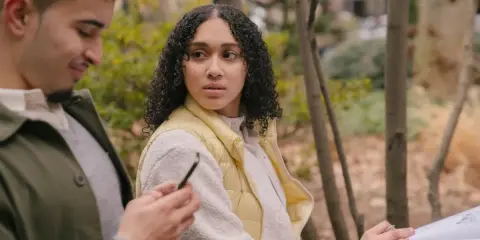 Un homme qui trompe et ment, regardant son téléphone, tandis qu'une femme se méfie, assise à côté de lui dans un parc.