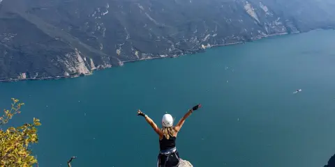Kobieta z rękami w górze na szczycie klifu, podziwiająca jezioro Garda po przejściu via ferrata.