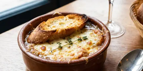 Un cuenco de sopa de cebolla gratinada con pan tostado y queso derretido, adornado con tomillo. Una copa de vino tinto y una cuchara completan la escena.