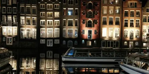 Edifici illuminati e riflessi sull'acqua, un battello turistico naviga tranquillo. Magica atmosfera di Amsterdam by night.