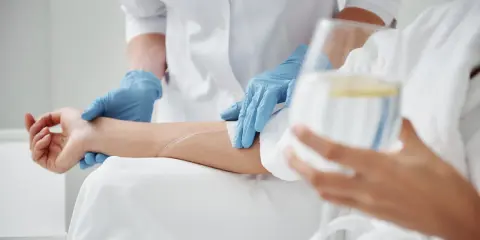 Pielęgniarka w niebieskich rękawiczkach zakłada wenflon do ręki pacjenta, przygotowując kroplówkę.