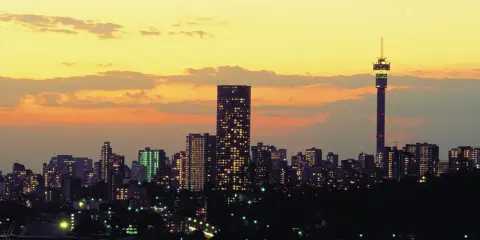 Stadssilhuett i Johannesburg, Sydafrika, med upplysta skyskrapor och ett högt torn mot en gul och orange solnedgångshimmel.
