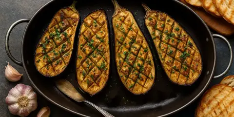 Quatre moitiés d'aubergine au four, grillées et parsemées de persil frais, dans un plat ovale avec de l'ail et des toasts grillés à côté.