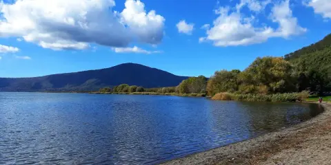 Spiaggia ghiaiosa e acque calme del lago di Vico, con montagne verdi e cielo azzurro punteggiato di nuvole bianche.