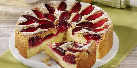 Szybkie ciasto ze śliwkami na oleju, posypane cukrem pudrem, z kawałkiem na łopatce. Obok miseczka z bitą śmietaną.