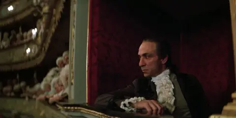 Scena z filmu o Mozarcie. Mężczyzna w stroju z epoki, w loży teatralnej, z widownią w tle.