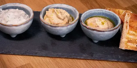Tre skålar med kall mat, troligen dippsåser eller röror, serveras med rostat bröd på en svart stenplatta.