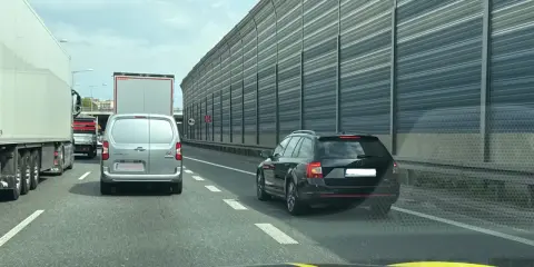 Korek na autostradzie. Samochody osobowe i ciężarowe stoją w rzędzie. W tle widać ekrany akustyczne. Jazda na suwak przepis to podstawa.