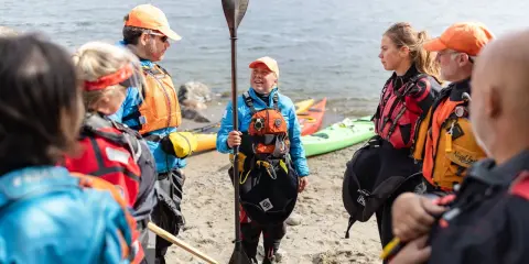 Grupp samlad på stranden, redo för paddling. En instruktör visar paddelteknik. Kajaker ligger i bakgrunden.