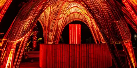 Struttura in bambù illuminata di rosso, con un bancone e un'installazione luminosa centrale. Atmosfera da house techno.