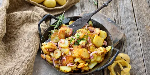 Bratkartoffeln, die vorher gekocht wurden, in einer Pfanne mit Speck und Kräutern garniert. Daneben liegen rohe Kartoffeln in einem Jutesack.