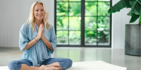Kvinna i blå kläder sitter i lotusställning på en matta, med händerna i bön. Hon utstrålar lugn, redo för en stunds shaking meditation.