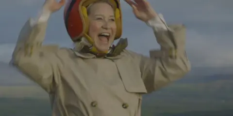 Glad Trine Dyrholm i pilotmössa och trenchcoat vinkar glatt. En scen från filmer med Trine Dyrholm.