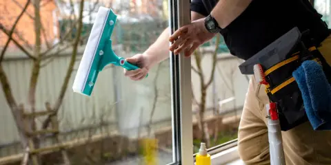 Osoba myje okno zieloną myjką, obok stoi żółty spray.