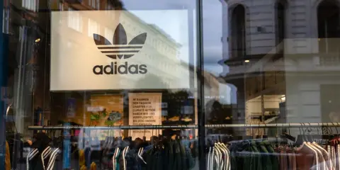 Witryna sklepu z logo adidas, marki pochodzącej z Niemiec. Wieszaki z ubraniami, w tym kurtki i bluzy z charakterystycznymi trzema paskami.