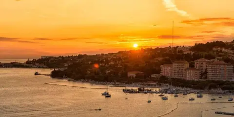 Tramonto dorato sul mare in Slovenia, con un porto pieno di barche e una costa verdeggiante. Immagina i bambini che giocano felici sulla spiaggia.
