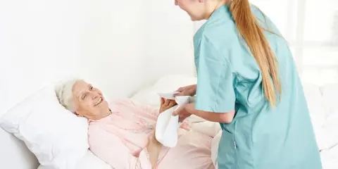 Opiekunka delikatnie myje krocze starszej pacjentce, która uśmiecha się z wdzięcznością.