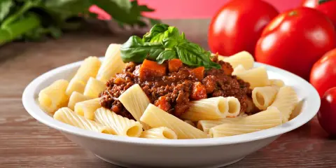 Pyszny makaron włoski rigatoni z sosem bolognese i świeżą bazylią, obok dojrzałe pomidory.