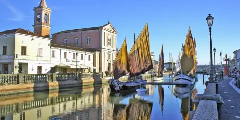 Traditional sailboats with colorful sails line a canal in Cesenatico, Italy, with buildings and a church reflected in the water.