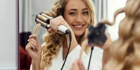 Uśmiechnięta blondynka w szlafroku tworzy piękne fale na włosach za pomocą karbownicy.