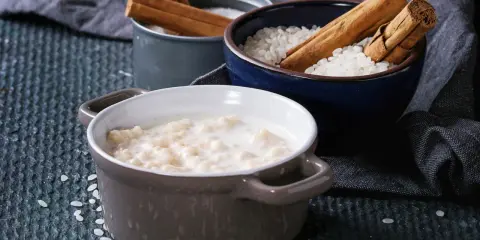 Gotowy ryż na mleku w miseczce, obok miseczki z ryżem i laskami cynamonu.