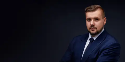 Mężczyzna w granatowym garniturze i krawacie, z zarostem, na ciemnym tle.