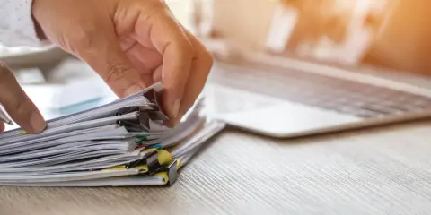 Dłonie przeglądają stos dokumentów, szukając odpowiedzi na pytanie, jak sprawdzić czy wpłynął pozew do sądu. Obok leży laptop.