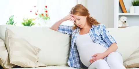 Zmęczona kobieta w ciąży, 26 tydzień, który miesiąc? Siedzi na kanapie, trzymając się za głowę.