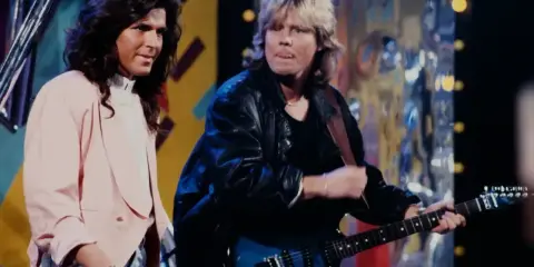 Dwóch muzyków na scenie, jeden gra na gitarze, drugi śpiewa. Ich styl przypomina teledysk do piosenki "tekst piosenki zawsze tam gdzie ty".
