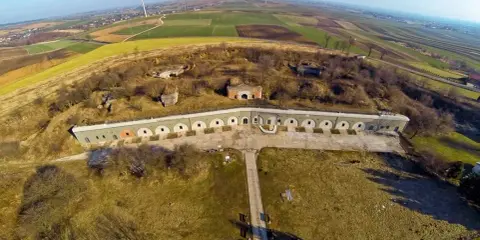 Widok z lotu ptaka na Twierdzę Przemyśl, z charakterystycznymi łukowymi oknami i otaczającymi ją polami.