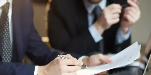 Dwóch mężczyzn w garniturach analizuje dokumenty. Jeden z nich wskazuje piórem na umowę, która może być naganą dla pracownika.
