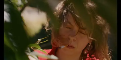 En närbild på Nina Widerberg med en cigarett i munnen, omgiven av gröna blad.