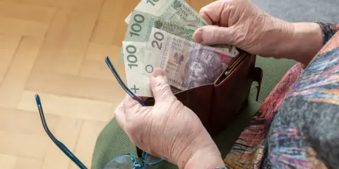 Dłonie starszej osoby wyciągają pieniądze z portfela. Widać banknoty 100 zł i 20 zł, być może to oczekiwana trzynasta emerytura.