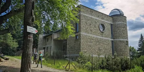 Kamienny budynek obserwatorium na szczycie Lubomir. Dwoje turystów podziwia widok, a obok drzewa i szlak PTTK.