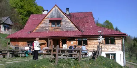 Schronisko pod Bereśnikiem, drewniany budynek z czerwoną dachem, otoczony zielenią. Kilka osób odpoczywa na ławkach przed schroniskiem.