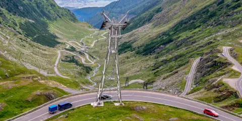 Kręte drogi w Rumunii wiją się przez zielone góry. Widać samochody i przyczepkę, a w oddali słup energetyczny.