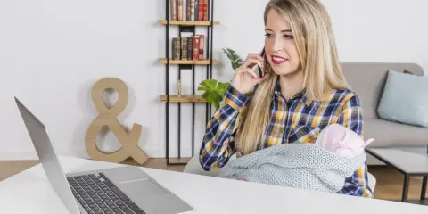 Kobieta z dzieckiem na rękach rozmawia przez telefon, pracując przy laptopie. To przykład pracy z orzeczeniem o niepełnosprawności, pokazujący elastyczność i możliwości.