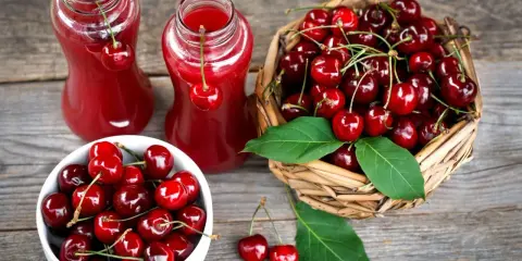 Dwa szklane dzbanki z rubinowym sokiem z czereśni, obok koszyk pełen świeżych owoców i biała miseczka z czereśniami.