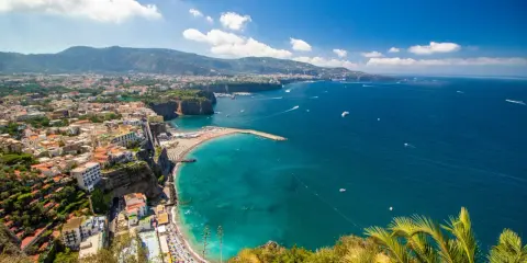 Vista panoramica della costa con il **scoglio del cavaliere** che emerge dal mare turchese, spiagge affollate e case aggrappate alla scogliera.
