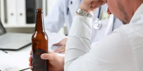 Mężczyzna trzyma butelkę piwa, siedząc przed lekarzem. Rozmowa sugeruje skierowanie na detoks alkoholowy.