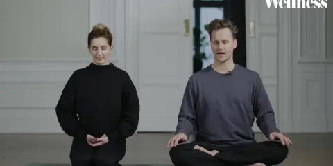 Två personer sitter på en yogamatta och mediterar. En kvinna i svart tröja och en man i grå tröja. Ta ett ögonblick att stanna upp.