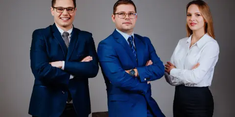 Trzy osoby w garniturach i białej koszuli, stojące na szarym tle, z rękami skrzyżowanymi na piersi.