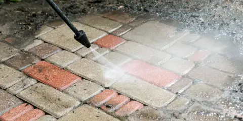 Czyszczenie kostki brukowej myjką ciśnieniową. Woda usuwa brud z czerwonych i szarych płyt.
