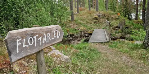 Träskylt med texten "Flottarleden Munkedal" pekar mot en liten träbro över en bäck i en skogsmiljö.