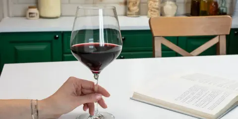 Dłoń trzyma kieliszek czerwonego wina, obok leży otwarta książka. Idealna lampka wina, by zatopić się w lekturze.