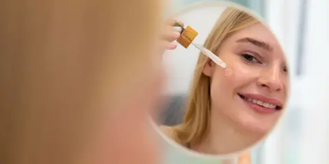 Mujer rubia sonríe mientras se aplica aceite facial con gotero en la mejilla. Un paso esencial en su rutina de cuidado de la piel, ya sea aceite facial antes o después de la crema.
