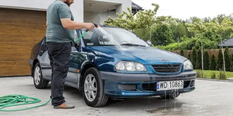Mężczyzna myje niebieskie auto na posesji, używając węża ogrodowego. Woda spływa po karoserii i tworzy kałuże na podjeździe.