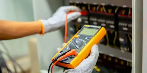 Elektryk w rękawicach wykonuje pomiar instalacji elektrycznej za pomocą żółtego multimetru.