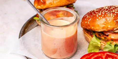 Ein Glas mit cremiger, rosafarbener beste burger sauce steht neben zwei saftigen Burgern. Ein Löffel ragt aus dem Glas.